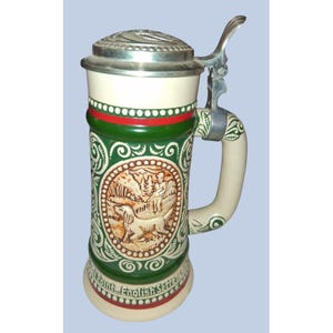 Puede incluir: Una jarra de cerveza de cerámica con tapa metálica abatible y asa. El cuerpo es de color crema con bandas verdes y rojas, mostrando una ilustración en relieve de una persona y un perro con el texto "At Point... English Setter" en la base.