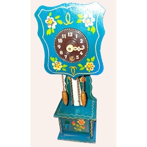 May include: A vintage, hand-painted wooden cuckoo clock in shades of teal and blue. The clock face is brown with white numbers and gold hands. Floral accents in yellow and white decorate the clock's frame and base. Two decorative pinecone weights hang below.