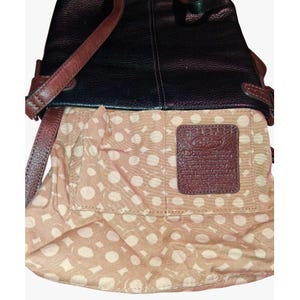 Puede incluir: Un bolso de cuero negro con correas y adornos de cuero marr&oacute;n. El interior est&aacute; forrado con una tela beige con un patr&oacute;n de lunares blancos. Un parche rectangular de cuero marr&oacute;n con texto en relieve est&aacute; cosido en el bolsillo interior.
