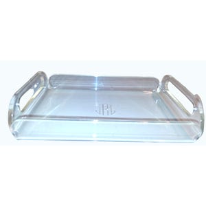 Puede incluir: Una bandeja de acrílico transparente con asas integradas. La bandeja tiene forma rectangular con esquinas redondeadas y un monograma en el centro. Las asas son lazos abiertos, y el diseño es minimalista y moderno.