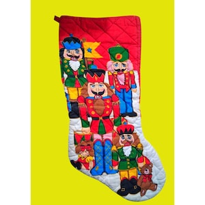 Puede incluir: Un calcetín navideño acolchado con la parte superior roja y la inferior blanca. El calcetín presenta soldados cascanueces aplicados en varios colores, incluyendo rojo, verde, azul y amarillo. También se incluye un osito de peluche.