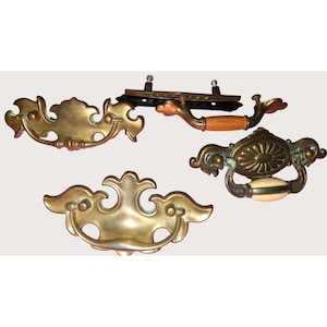 Könnte beinhalten: Vier antike Schubladengriffe und -griffe aus Messing. Die Griffe haben verzierte Designs, darunter Blumen- und Scrollmuster. Ein Griff hat einen Holzgriff. Die Hardware ist in verschiedenen Formen und Größen erhältlich, geeignet für die Möbelrestaurierung oder Vintage-Dekoration.