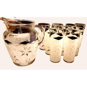 May include: A clear glass pitcher and eight matching tall drinking glasses, each decorated with a frosted white and silver snowflake pattern and a band of black diamond shapes near the top rim, creating a cohesive set for serving beverages.