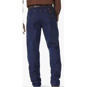 May include: Dark blue denim jeans with the Wrangler logo on the back pockets. The jeans have a classic fit and are worn with a brown belt. The jeans are a staple in Western wear.