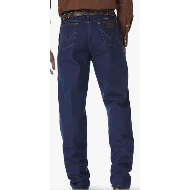 May include: Dark blue denim jeans with the Wrangler logo on the back pockets. The jeans have a classic fit and are worn with a brown belt. The jeans are a staple in Western wear.