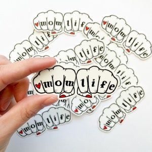 K&ouml;nnte beinhalten: Ein Satz wei&szlig;er Aufkleber mit einem roten Herzen und dem Text "mom life" in schwarzen Buchstaben, angeordnet in einem Kreis. Die Aufkleber sind so gestaltet, dass sie wie eine Faust machende Hand aussehen.