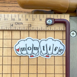 K&ouml;nnte beinhalten: Wei&szlig;er Aufkleber mit vier F&auml;usten, die das Wort "momlife" in schwarzen Buchstaben bilden. Ein kleines rotes Herz befindet sich in der ersten Faust.