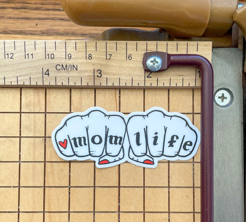 K&ouml;nnte beinhalten: Wei&szlig;er Aufkleber mit vier F&auml;usten, die das Wort "momlife" in schwarzen Buchstaben bilden. Ein kleines rotes Herz befindet sich in der ersten Faust.