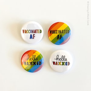 May include: Four round buttons with text. Two buttons have the text "VACCINATED AF" in rainbow colors and black. The other two buttons have the text "hello VAXXED" in rainbow colors and black. The buttons are white and rainbow colored.