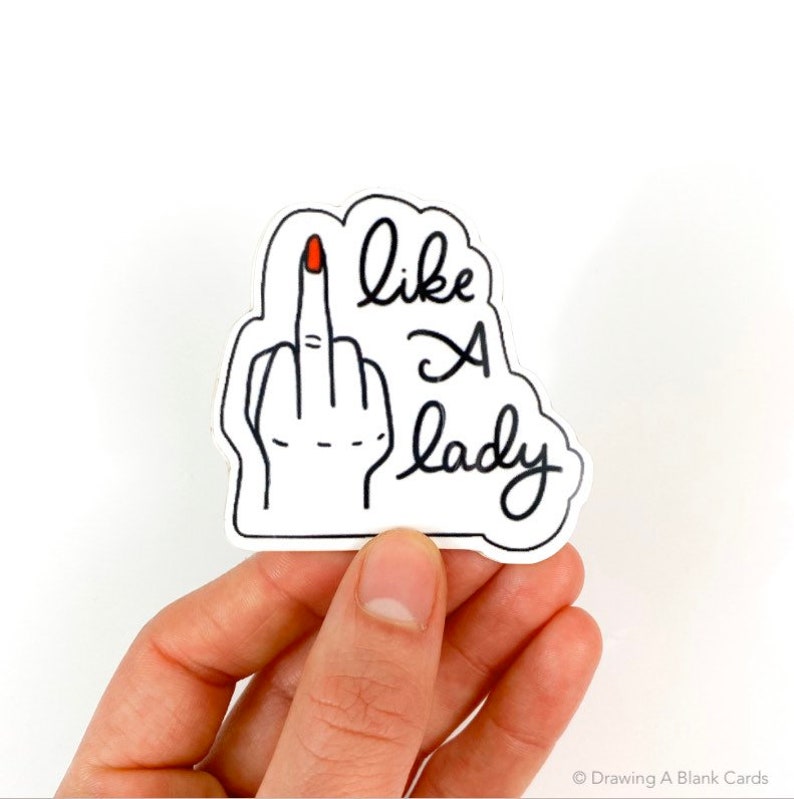 May include: White sticker with a black line drawing of a hand giving the middle finger. The middle finger has a red nail. The text "like a lady" is written in black cursive.