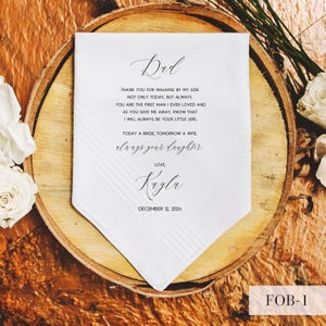 May include: A white handkerchief with a printed message for a father from his daughter on her wedding day. The text includes the words "Dad", "Love, Kayla", and the date "December 12, 2024". The handkerchief is on a wooden surface.