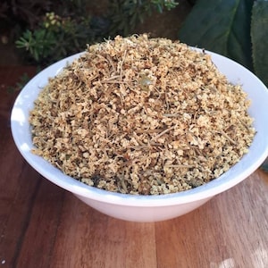 May include: A white bowl brimming with dried elderflowers, pale brown in colour. The bowl rests on a wooden surface, with green foliage and pink flowers visible in the background. The elderflowers look freshly picked.
