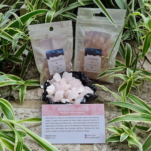 May include: Two small bags of pink rose quartz crystal chips, labelled "Evies House Crystal Chips 100g". A small bowl filled with rose quartz chips sits in front of the bags. A card with the text "Rose Quartz Femininity Love Kindness" and information about the stone's properties is below the bowl.
