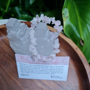 May include: A pink rose quartz chip bracelet displayed on a white crystal cluster. The bracelet is made of small, irregularly shaped rose quartz chips. The text on the card reads "Rose Quartz - Femininity - Love - Kindness".
