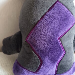 Puede incluir: Un juguete de peluche con forma de criatura con cuerpo gris y detalles morados. El juguete tiene un dise&ntilde;o de rayo en la espalda.