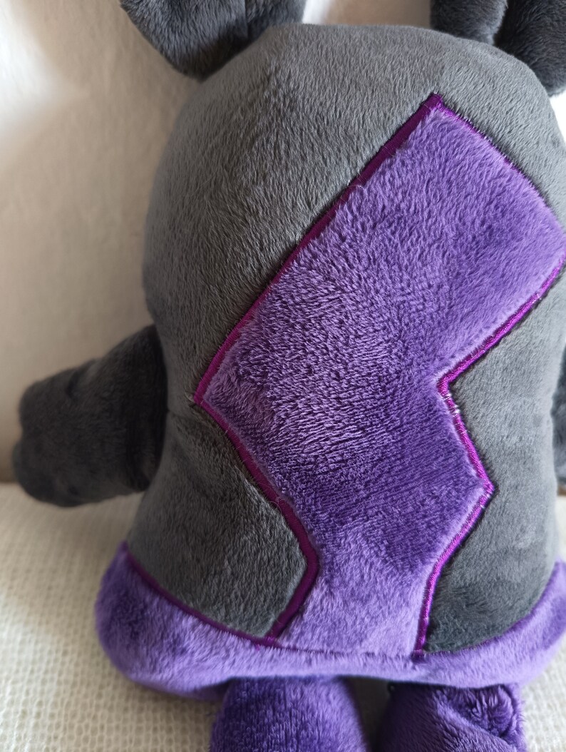 Puede incluir: Un juguete de peluche con forma de criatura con cuerpo gris y detalles morados. El juguete tiene un dise&ntilde;o de rayo en la espalda.