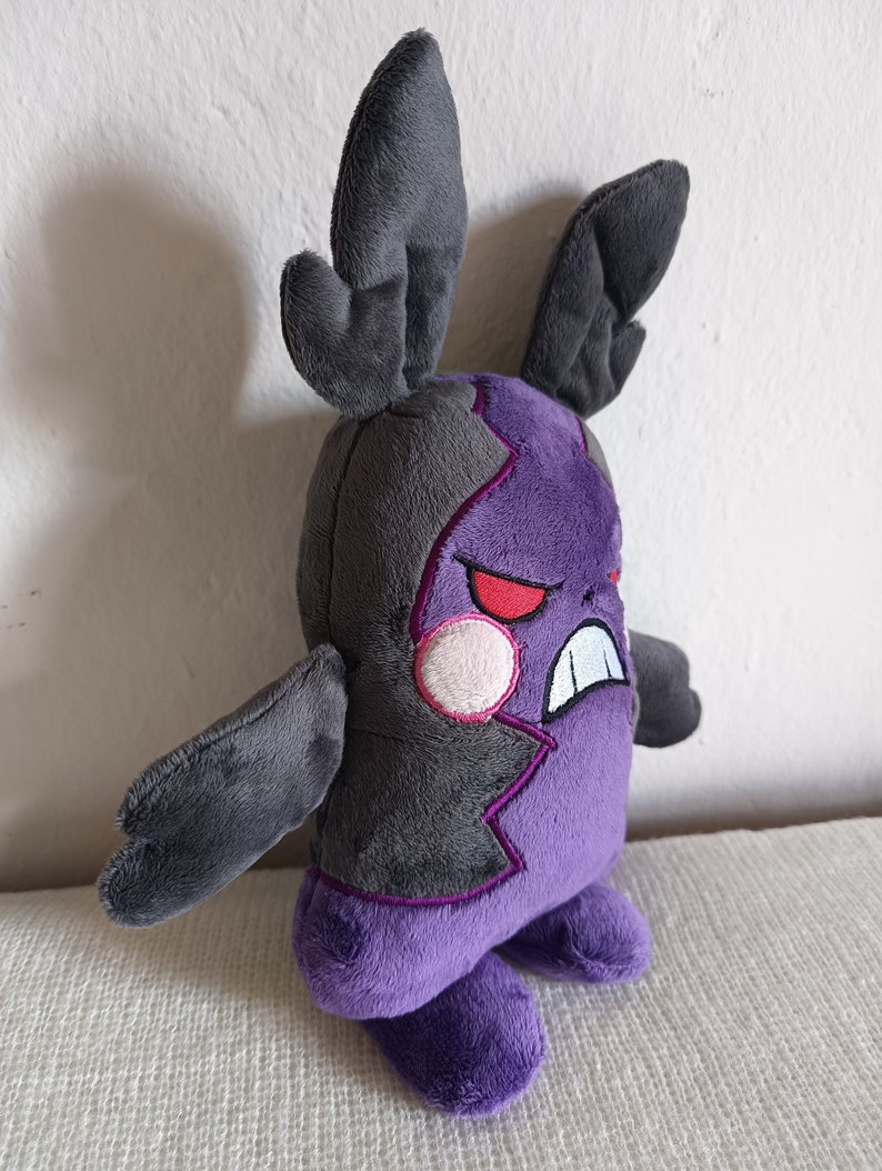 Puede incluir: Juguete de peluche morado y gris con orejas grandes y una cara fruncida. El juguete tiene un dise&ntilde;o morado dentado en su cuerpo y un c&iacute;rculo rosa en su cara.