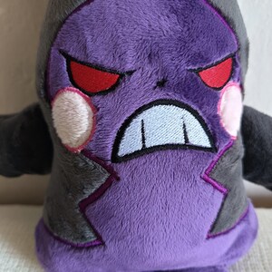 Puede incluir: Juguete de peluche morado con detalles negros y ojos rojos. El juguete tiene una expresi&oacute;n de enfado con dientes blancos.