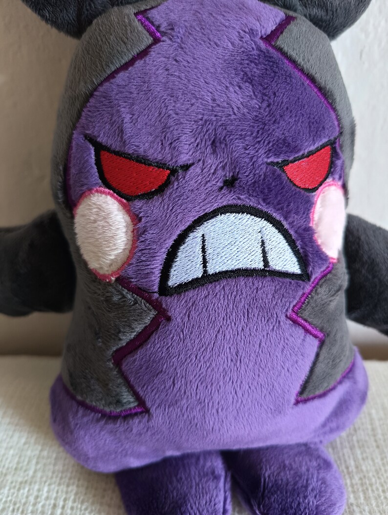 Puede incluir: Juguete de peluche morado con detalles negros y ojos rojos. El juguete tiene una expresi&oacute;n de enfado con dientes blancos.
