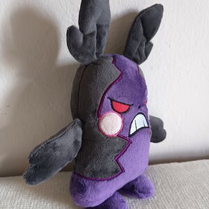 Puede incluir: Juguete de peluche morado y gris con orejas grandes y una expresi&oacute;n amenazante. El juguete tiene un dise&ntilde;o morado y gris dentado y ojos rojos.