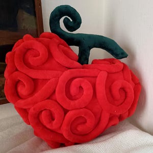 Könnte beinhalten: Ein rotes, plüschiges, fruchtförmiges Kissen mit einem grünen Stiel. Das Kissen hat eine strukturierte Oberfläche mit wirbelnden Mustern. Der grüne Stiel ist nach oben gebogen. Das Kissen liegt auf einer weißen Oberfläche.