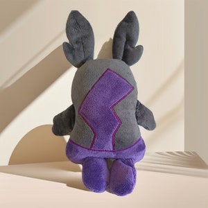 Puede incluir: Un juguete de peluche con forma de criatura morada y gris con orejas grandes. La criatura tiene un dise&ntilde;o de rayo morado en el pecho.