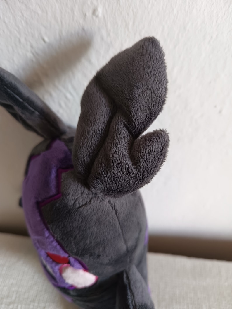 Puede incluir: Un juguete de peluche de color gris y morado. El juguete tiene orejas grandes y ca&iacute;das y un cuerpo peque&ntilde;o y redondo. El juguete est&aacute; hecho de tela suave y peluda.