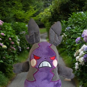 Puede incluir: Juguete de peluche morado y gris con una expresi&oacute;n amenazante. El juguete tiene orejas grandes y una boca ancha con dientes afilados. El juguete est&aacute; de pie en un camino en un jard&iacute;n.