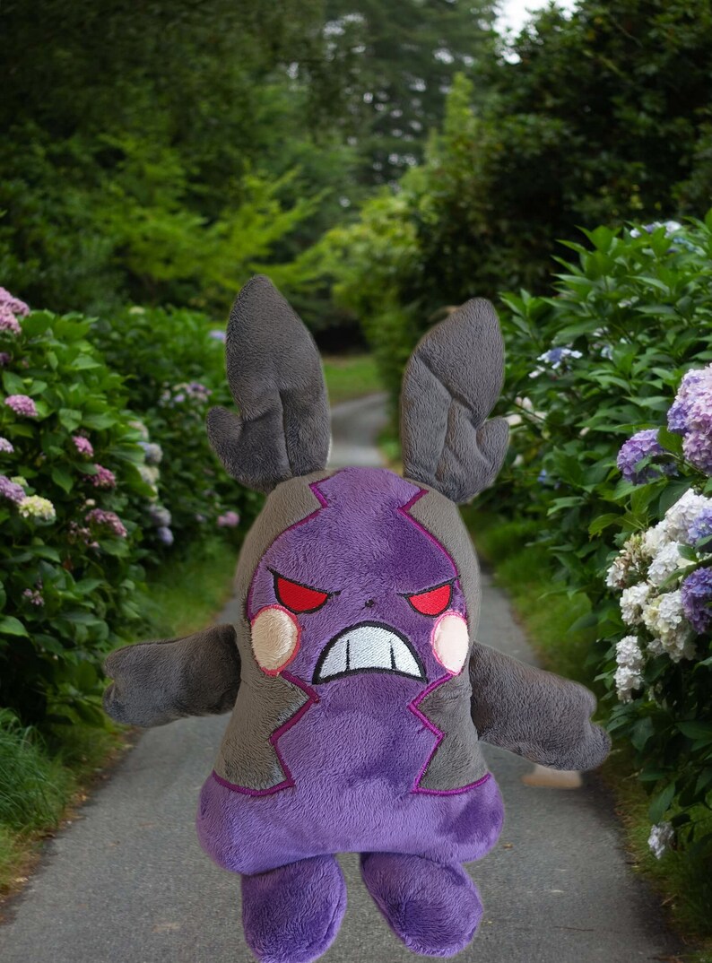 Puede incluir: Juguete de peluche morado y gris con una expresi&oacute;n amenazante. El juguete tiene orejas grandes y una boca ancha con dientes afilados. El juguete est&aacute; de pie en un camino en un jard&iacute;n.