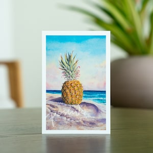 May include: A watercolor painting of a pineapple on a beach with blue water and a pink sky. The pineapple is in the center of the image and is surrounded by sand. The painting is signed "pedluky" in the bottom right corner.