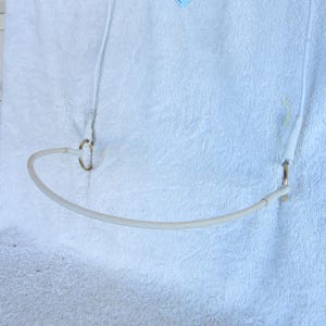 Puede incluir: Un tubo blanco, curvo y flexible con un pequeño anillo dorado en cada extremo. El tubo está conectado a dos cordones blancos. El artículo está sobre una superficie blanca y texturizada.