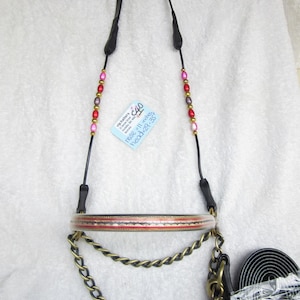 Puede incluir: Un cabezal de caballo de cuero negro con una boquilla trenzada roja y dorada, una cadena de latón y cuentas rosas y rojas. El cabezal está etiquetado como "Vig Hatters" y tiene el texto "Nose 11 inches Head 22-25".