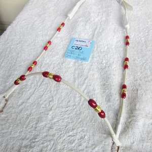 Puede incluir: Un cabezada de cuero blanco con cuentas rojas y doradas. La cabezada está sobre una superficie blanca y esponjosa. Una nota azul pegada a la cabezada dice "vip.halters C20 nose - 11 1/2 sides - 16 (29 - 35 gall)"