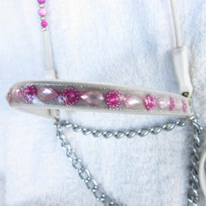 Puede incluir: Un cabezal de caballo de cuero blanco con diamantes de imitación rosas y transparentes. El cabezal tiene una cadena plateada.
