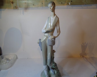 May include: A porcelain figurine of a person standing with a bag over their shoulder. The figurine is painted in shades of white, grey, and brown. The person is wearing a white shirt, a brown waistcoat, and brown trousers. The figurine is standing on a base that is painted green.