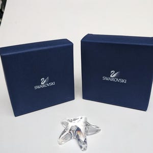 Op de afbeelding: Een helder kristallen zeesterfiguur ligt voor twee marineblauwe dozen. De dozen hebben het Swarovski-logo in wit. De zeester heeft vijf punten en reflecteert licht. De dozen zijn effen van kleur.
