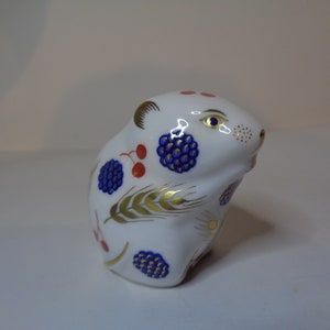 Puede incluir: Figura de cerámica blanca de un hámster, decorada con bayas azules, cerezas rojas y trigo dorado. El hámster tiene detalles dorados en las orejas y los ojos, y es un objeto decorativo.