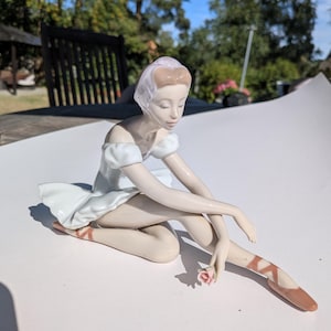 Puede incluir: Una figura de porcelana de una bailarina sentada con un vestido blanco de mangas abullonadas. La figura tiene piel clara, cabello castaño recogido en un moño y sostiene una pequeña rosa rosada. La bailarina lleva zapatillas de ballet marrones.