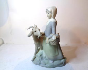 Könnte beinhalten: Eine Porzellanfigur einer jungen Frau in einem grauen Kleid und einer weißen Haube, die einen Korb hält und mit einer weißen Ziege geht.