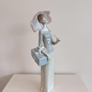 May include: A porcelain figurine of a woman holding a parasol and a gift box. The woman wears a grey top and a long, cream-coloured skirt. The parasol is white with a pink trim. The gift box is light blue with white polka dots.