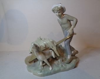 May include: A porcelain figurine of a boy in a straw hat pushing a wheelbarrow with a dog standing beside it. The wheelbarrow is filled with hay and the boy is wearing a brown shirt and blue pants.