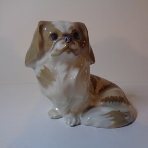 May include: A porcelain figurine of a brown and white Pekingese dog sitting with its head tilted up. The dog has a white chest and a brown and white tail.