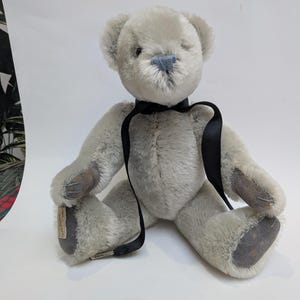 May include: A light gray teddy bear with a black ribbon around its neck. The bear has a blue nose and dark gray paw pads. The bear is sitting on a white surface.