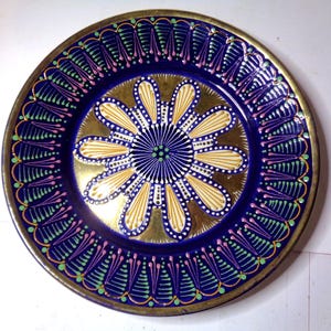 Könnte beinhalten: Ein dekorativer Keramikteller mit blauem Hintergrund und einem goldenen Blumendesign. Die Blume hat eine weiße Mitte und gelbe Blütenblätter mit weißen Punkten. Der Teller ist von einem Rand aus grünen und rosa Wirbeln umgeben.