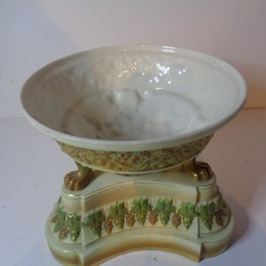 May include: A white ceramic bowl with gold and green floral designs sits on a pedestal with three gold lion paw feet. The pedestal is decorated with green leaves and gold grapes.
