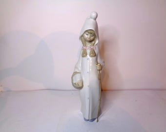May include: A porcelain figurine of a girl wearing a white hooded robe and holding a stick. She has a pink ribbon around her neck and is standing on a white base.