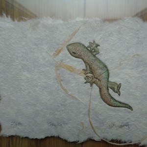 May include: A green and brown gecko is climbing a thin, light brown branch. The gecko is depicted in a watercolour style on a white textured paper. The paper has a rough, handmade look. The image is signed "Gecko" and numbered "2/50".