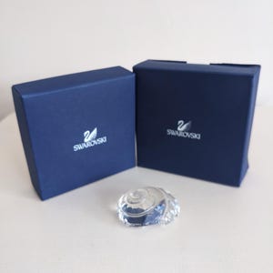 Może przedstawiać: Przezroczysta kryształowa figurka muszli leży na białej powierzchni, obok dwóch granatowych pudełek. Pudełka mają białe logo Swarovski. Muszla jest szczegółowa i odbija światło.