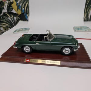 Könnte beinhalten: Ein dunkelgrünes 1963 MGB Roadster Modellauto mit schwarzem Innenraum und silbernen Rädern. Das Fahrzeug ist auf einem rechteckigen Holzsockel montiert, der eine goldfarbene Plakette mit dem Text CORGI 1963 MGB ROADSTER trägt.