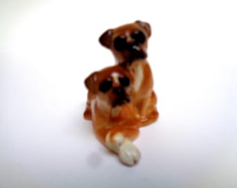 May include: Two small ceramic figurines of brown and white boxer dogs. The dogs are in a playful pose, with one dog sitting and the other lying down.